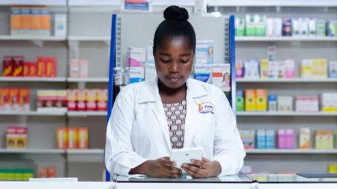 A pharmacist checking a phone behind a counter, shelves of medicines behind her